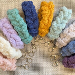 May include: A set of ten colorful crocheted keychains with silver key rings. The keychains are made of a soft, fluffy yarn and are arranged in a semi-circle.