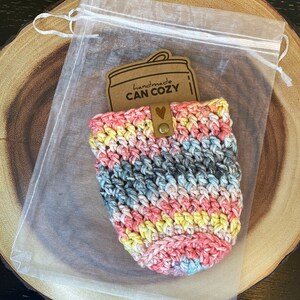 May include: A crocheted can cozy in a rainbow of colors, pink, blue, yellow, and gray. The cozy is in a clear organza bag with a brown paper tag that says "handmade CAN COZY".