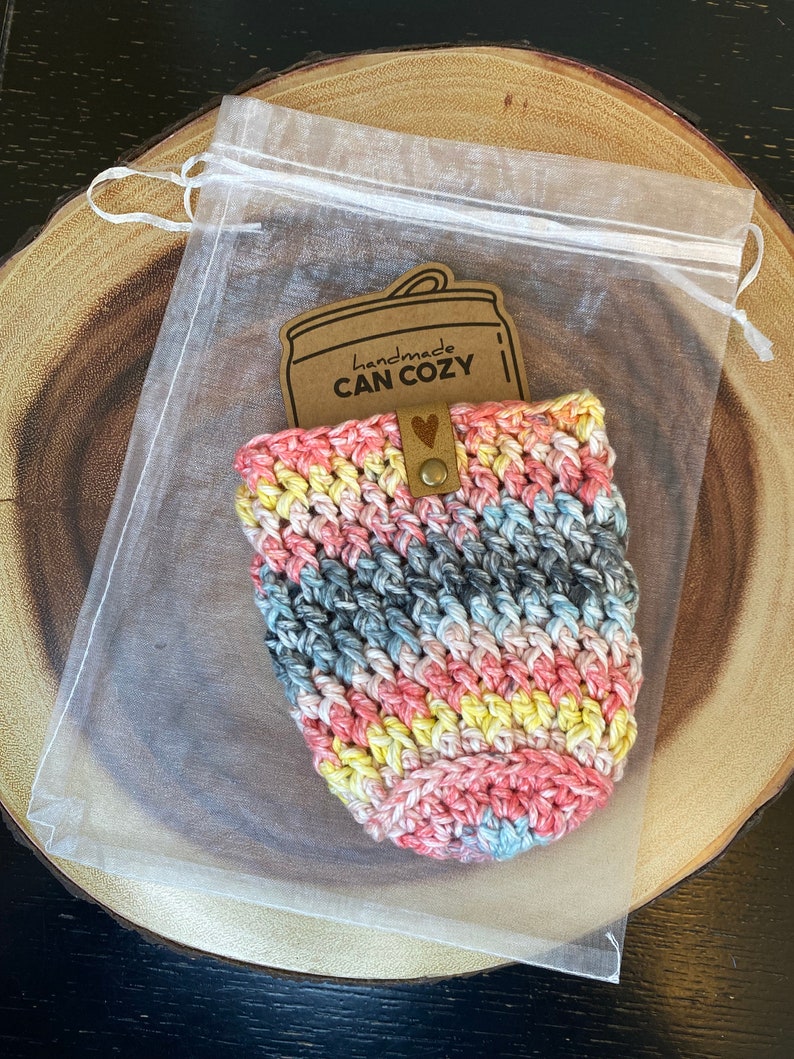 May include: A crocheted can cozy in a rainbow of colors, pink, blue, yellow, and gray. The cozy is in a clear organza bag with a brown paper tag that says "handmade CAN COZY".
