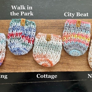 May include: Five crocheted cup cozies in different color patterns. The cozies are labeled "Walk in the Park", "City Beat", "Sunsetting", "Cottage", and "Neutral".