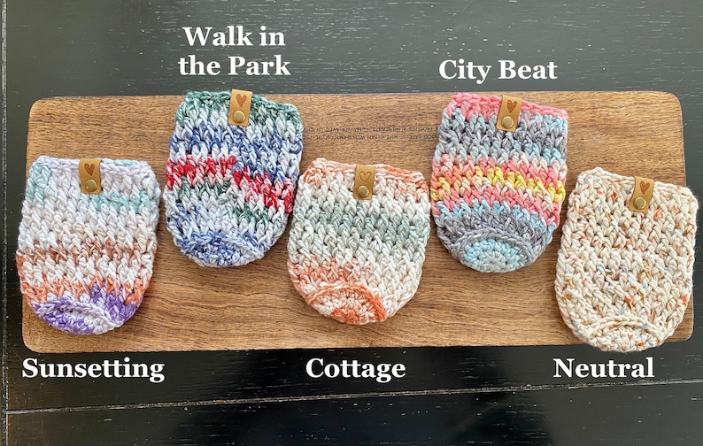 May include: Five crocheted cup cozies in different color patterns. The cozies are labeled "Walk in the Park", "City Beat", "Sunsetting", "Cottage", and "Neutral".