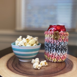 May include: A crocheted can cozy in pink, yellow, and gray with a brown heart-shaped button. A bowl of popcorn and a can of soda are on a wooden slice.
