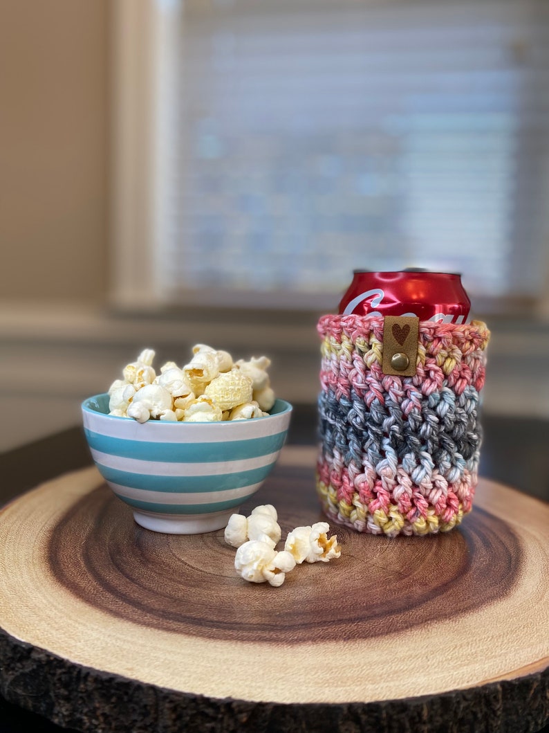 May include: A crocheted can cozy in pink, yellow, and gray with a brown heart-shaped button. A bowl of popcorn and a can of soda are on a wooden slice.