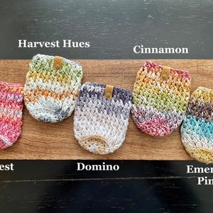 May include: Five crocheted cup cozies in different color combinations, including Harvest Hues, Cinnamon, Domino, and Emerald Pine. Each cozy has a brown leather tag with a heart.