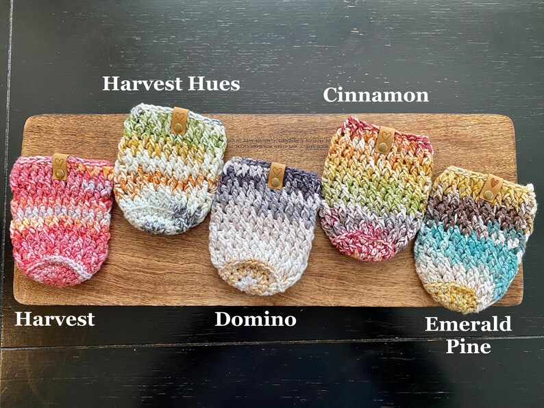 May include: Five crocheted cup cozies in different color combinations, including Harvest Hues, Cinnamon, Domino, and Emerald Pine. Each cozy has a brown leather tag with a heart.