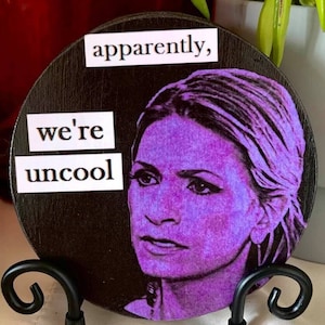 May include: A round wooden coaster with a purple image of a woman's face and the text "apparently, we're uncool" in white.