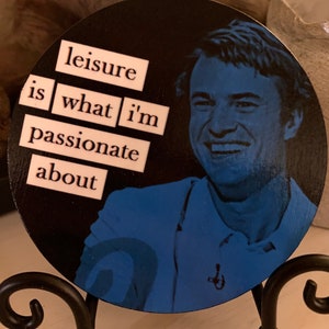 May include: A round wooden coaster with a black background and a blue and white image of a man smiling. The words "leisure is what i'm passionate about" are written in white letters on the coaster.