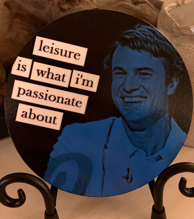 May include: A round wooden coaster with a black background and a blue and white image of a man smiling. The words "leisure is what i'm passionate about" are written in white letters on the coaster.