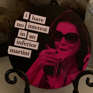 May include: A round wooden coaster with a black background and a pink image of a woman wearing sunglasses and holding a martini glass. The text "i have no interest in an inferior martini" is printed in pink on the coaster.