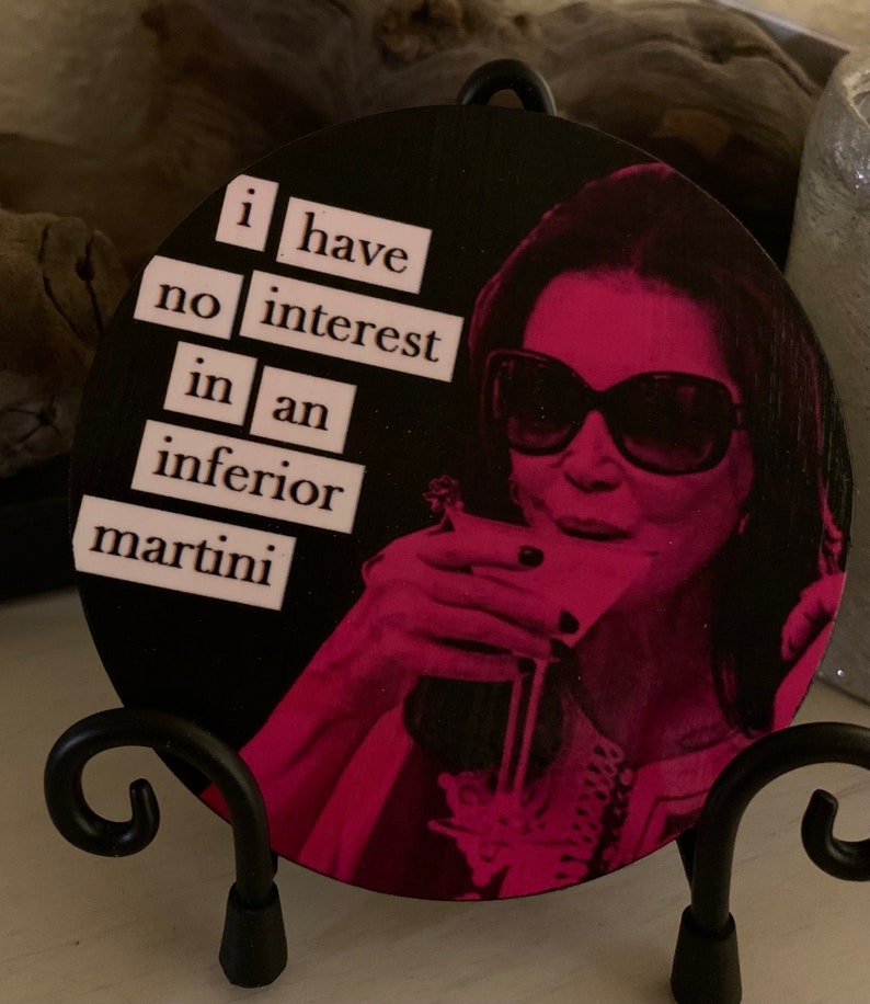 May include: A round wooden coaster with a black background and a pink image of a woman wearing sunglasses and holding a martini glass. The text "i have no interest in an inferior martini" is printed in pink on the coaster.