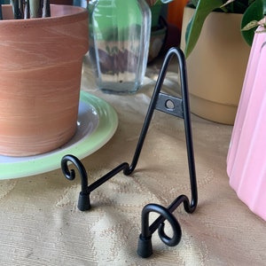 May include: Black metal display stand with two curled feet. The stand is designed to hold a plate or other flat object.