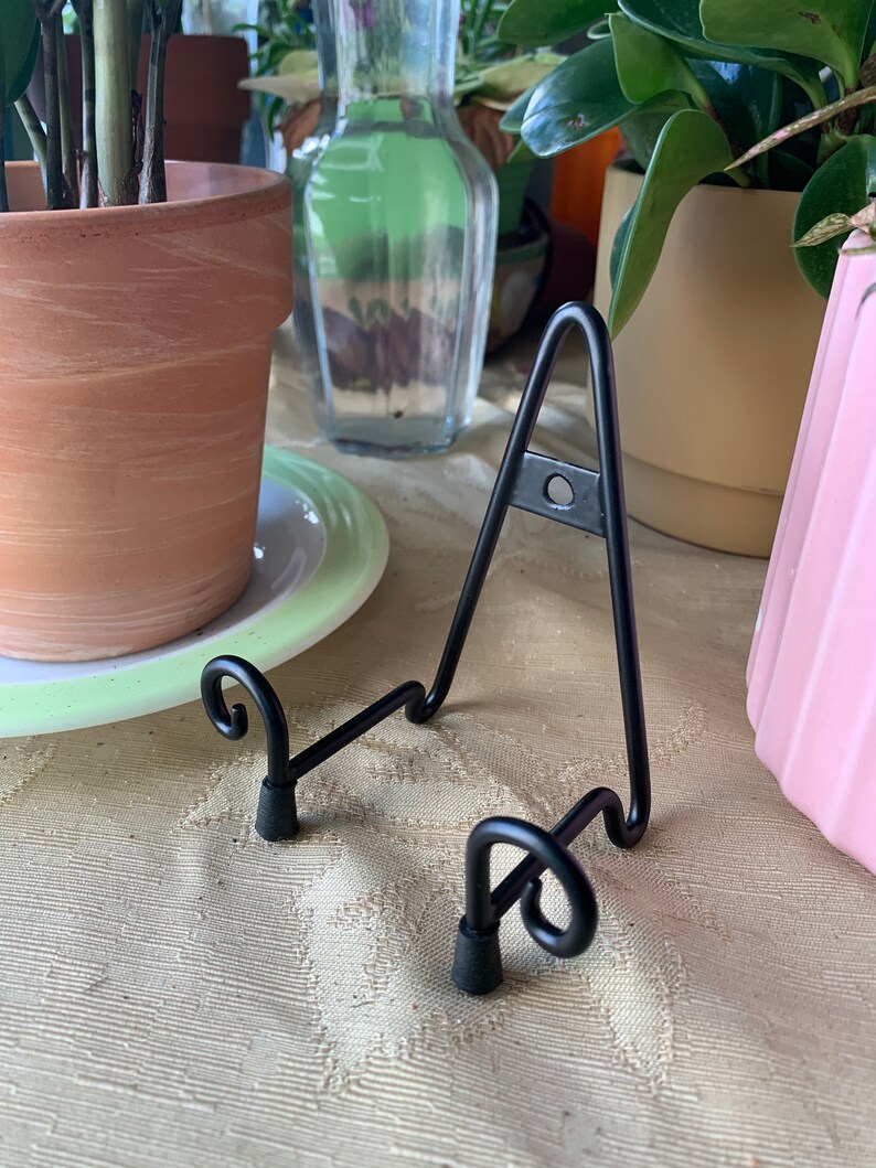 May include: Black metal display stand with two curled feet. The stand is designed to hold a plate or other flat object.