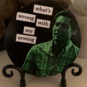 May include: A black and white round wooden coaster with a green image of a man with the text "what's wrong with my sewing" printed on it. The coaster is on a black metal stand.