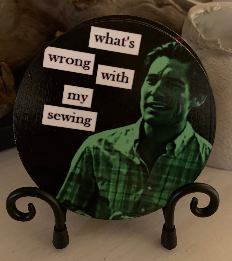 May include: A black and white round wooden coaster with a green image of a man with the text "what's wrong with my sewing" printed on it. The coaster is on a black metal stand.