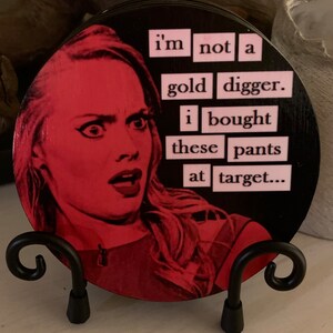 May include: A round coaster with a black background and a pink and white image of a woman with the text "i'm not a gold digger. i bought these pants at target..." printed on it.