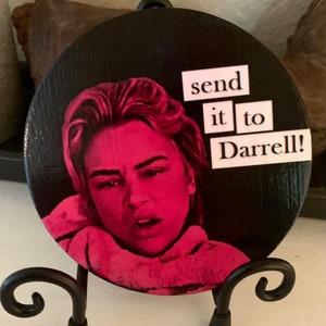 May include: A round wooden sign with a pink image of a woman with a distressed expression. The sign has white text that reads "send it to Darrell!"