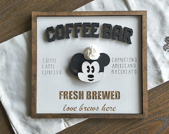 May include: Wooden coffee bar sign with a white background and a dark brown frame. The sign features the words "COFFEE BAR" in black letters, a Mickey Mouse design, and the words "FRESH BREWED" and "love brews here".