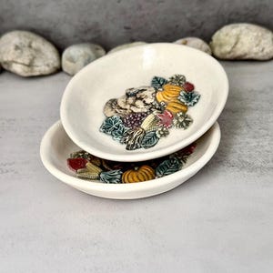 Peut inclure: Deux petits plats en céramique blanc cassé empilés. Chaque plat présente un motif en relief coloré d'éléments de la récolte d'automne, dont des citrouilles, du maïs et des raisins. Le fond est gris neutre.