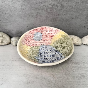 May include: A small, off-white ceramic bowl with a rainbow-colored interior. The bowl's interior is decorated with the repeated text "Chag Sameach" in various colors. The bowl is set against a gray background.