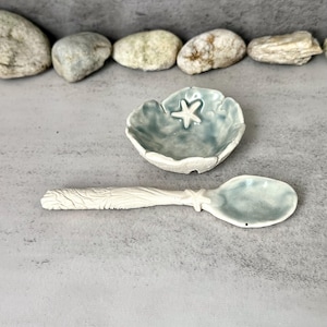 Peut inclure: Un ensemble de cuillère et de petit bol en céramique sur le thème marin. La cuillère et le bol sont bleu clair et blancs, avec un détail d'étoile de mer sur le bol. La cuillère a un manche texturé. L'ensemble est présenté sur une surface grise.