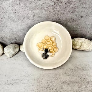 Puede incluir: Un pequeño plato de cerámica blanquecina con un diseño en relieve de un panal y una abeja. La abeja es negra y blanca con alas amarillas, y el panal es de color amarillo claro. El plato es redondo.