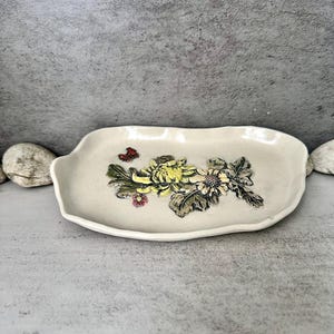 Peut inclure: Un plateau rectangulaire en céramique blanc cassé avec un motif floral. Le plateau présente un groupe de fleurs jaunes, blanches et vertes, ainsi qu'un petit papillon rouge. Les bords du plateau sont légèrement ondulés.