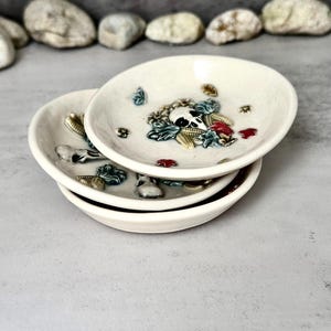 Peut inclure: Trois petites coupelles en céramique blanc cassé empilées. Chaque coupelle présente un motif de crâne et de fleurs avec des accents bleus, verts et rouges. Les coupelles sont rondes avec un bord légèrement surélevé.