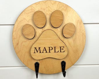 May include: A round, light-colored wooden paw print-shaped wall hanging with the name "MAPLE" engraved in the center. It features five raised paw pads and two black metal hooks below. The wall hanging is mounted on a white horizontal plank wall.