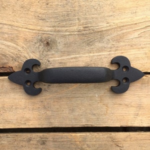 May include: Black cast iron door pull with a fleur-de-lis design. The pull has a smooth, black finish and is mounted on a wooden surface.