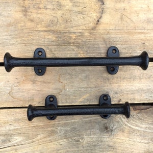 May include: Two black cast iron towel bars. Each bar has a cylindrical centre and rounded ends. The bars are mounted on rectangular plates with two screw holes. The bars are displayed on a light brown wooden surface.
