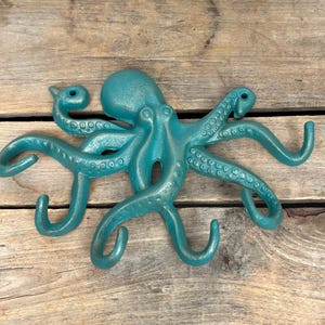 May include: A teal octopus wall hook made of metal. The octopus has eight tentacles, each ending in a hook. The hook is mounted on a wooden surface.