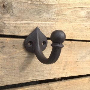 May include: A black cast iron hook with a rounded top and a pointed base. The hook is mounted on a wooden surface.