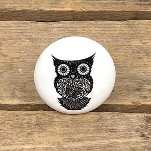 May include: A white, circular decorative knob with a detailed black owl design. The owl is illustrated with intricate patterns and a symmetrical arrangement, creating a striking contrast against the white background. The knob is likely made of ceramic or a similar material.