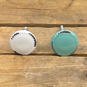 May include: Two round cabinet knobs. One is white with a distressed black edge, the other is turquoise with a distressed black edge. Both have silver stems and are set against a wooden background.