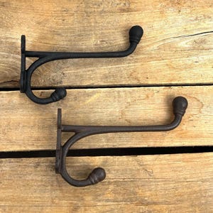 K&ouml;nnte beinhalten: Zwei schwarze Gusseisen-Kleiderhaken mit rustikalem Design. Jeder Haken hat einen gebogenen Arm, der in einer dekorativen Kugel endet. Die Haken sind auf einer Holzoberfl&auml;che montiert und zeigen ihren Vintage-Charme. Ideal f&uuml;r Eingangsbereiche oder Schlafzimmer.