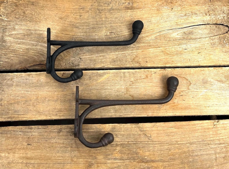 K&ouml;nnte beinhalten: Zwei schwarze Gusseisen-Kleiderhaken mit rustikalem Design. Jeder Haken hat einen gebogenen Arm, der in einer dekorativen Kugel endet. Die Haken sind auf einer Holzoberfl&auml;che montiert und zeigen ihren Vintage-Charme. Ideal f&uuml;r Eingangsbereiche oder Schlafzimmer.