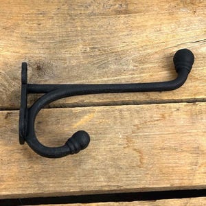 K&ouml;nnte beinhalten: Ein schwarzer Wandhaken aus Gusseisen mit geschwungenem Design. Der Haken ist auf einer Holzoberfl&auml;che montiert. Der Haken hat eine dekorative Kugel am Ende des gebogenen Arms und am Ende der Montageplatte.