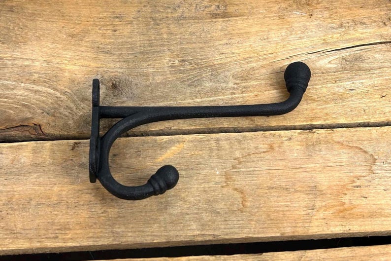 K&ouml;nnte beinhalten: Ein schwarzer Wandhaken aus Gusseisen mit geschwungenem Design. Der Haken ist auf einer Holzoberfl&auml;che montiert. Der Haken hat eine dekorative Kugel am Ende des gebogenen Arms und am Ende der Montageplatte.