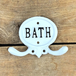 May include: White metal wall hook with the word "BATH" in black lettering. The hook has a decorative oval shape with two curved arms extending outwards. The hook is mounted on a wooden surface.