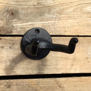 May include: A black cast iron wall hook with a circular base and a curved hook. The hook is mounted on a wooden surface. The hook is designed for hanging items. The hook is a dark matte black color.