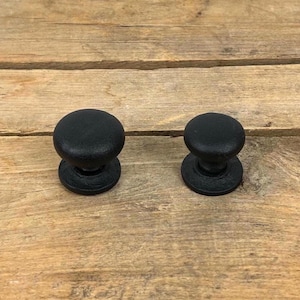 May include: Two black cabinet knobs with a rounded top and a circular base are displayed on a wooden surface. The knobs are made of a matte black material and are of different sizes. The wood grain is visible in the background.
