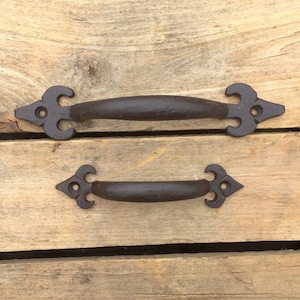 Peut inclure: Deux poignées de porte en fonte noire avec un motif décoratif de fleur de lys. Les poignées sont présentées sur un fond en bois.