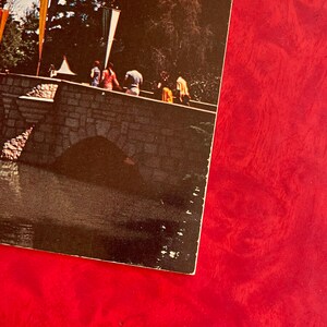 May include: A vintage postcard featuring a stone bridge with people walking across it. The bridge has a stone archway and is surrounded by trees and a small body of water. The postcard is faded and has a red background.