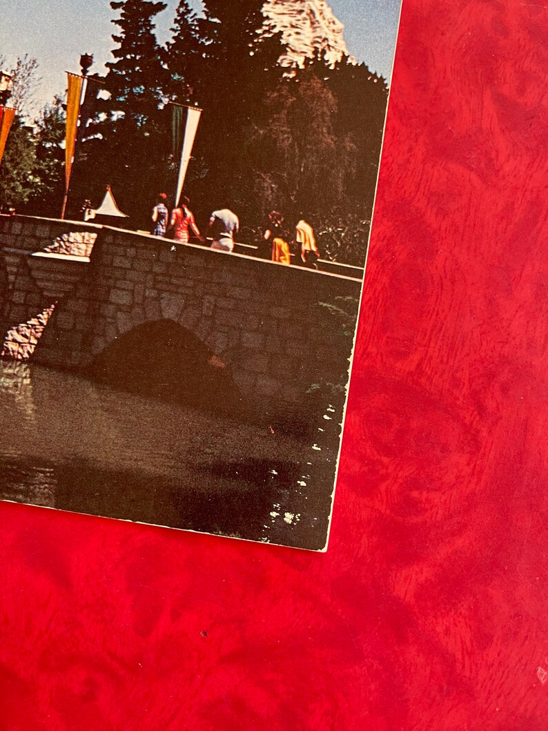 May include: A vintage postcard featuring a stone bridge with people walking across it. The bridge has a stone archway and is surrounded by trees and a small body of water. The postcard is faded and has a red background.