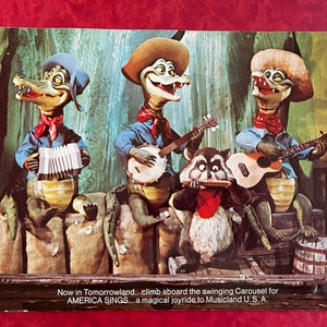 May include: A vintage postcard featuring a cartoon band of three alligators and a raccoon playing musical instruments. The alligators are wearing cowboy hats and overalls. The raccoon is wearing a red bandana. The text on the postcard reads: "Now in Tomorrowland...climb aboard the swinging Carousel for AMERICA SINGS...a magical joyride to Musicland U.S.A."