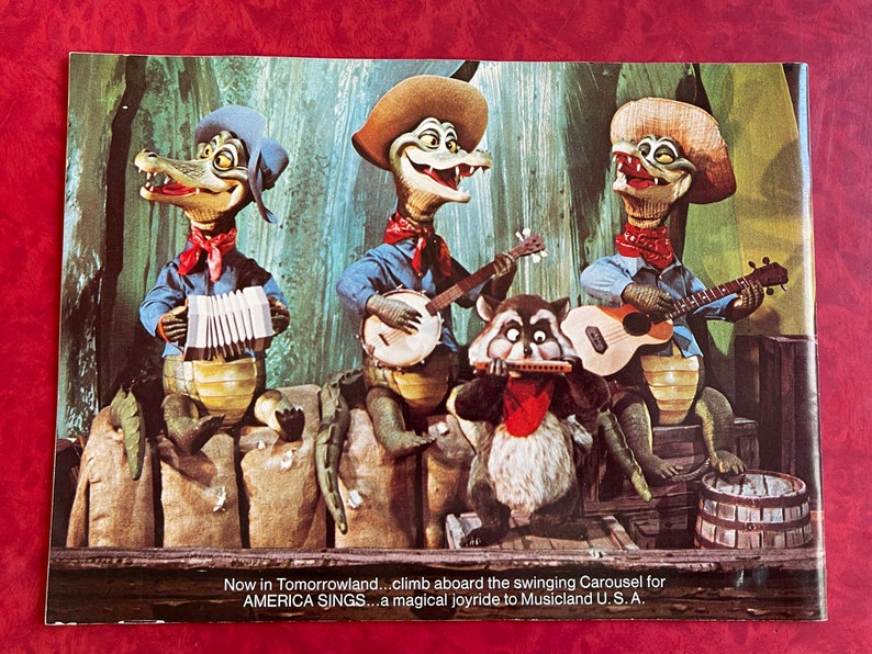 May include: A vintage postcard featuring a cartoon band of three alligators and a raccoon playing musical instruments. The alligators are wearing cowboy hats and overalls. The raccoon is wearing a red bandana. The text on the postcard reads: "Now in Tomorrowland...climb aboard the swinging Carousel for AMERICA SINGS...a magical joyride to Musicland U.S.A."