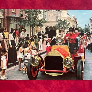 May include: Mickey Mouse in a red and black costume drives a vintage red car with gold accents.  The car is surrounded by people, including children, in a street setting.  The iconic Cinderella's Castle is visible in the background.