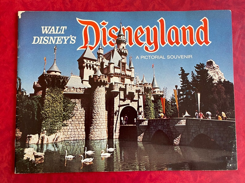 May include: A vintage book cover featuring a colorful illustration of Sleeping Beauty's castle at Disneyland. The text on the cover reads "Walt Disney's Disneyland A Pictorial Souvenir".