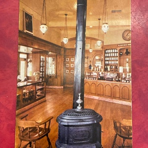 May include: A vintage interior shot of a shop with a wooden floor, wood panelling, and a black cast iron wood-burning stove. The shop has a counter with shelves and jars of product. There are several hanging light fixtures with glass globes.