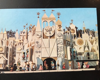 May include: A vintage postcard featuring a colorful, whimsical building with a clock face and a variety of architectural details. The building is surrounded by smaller buildings and trees. There are figures of people in front of the building.